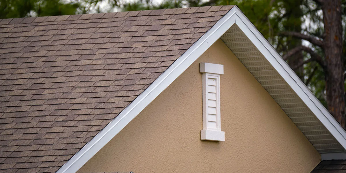 Close-up of a house roof with brown architectural shingles and part of a beige wall featuring a white attic vent. Green tree branches appear in the blurred background.
