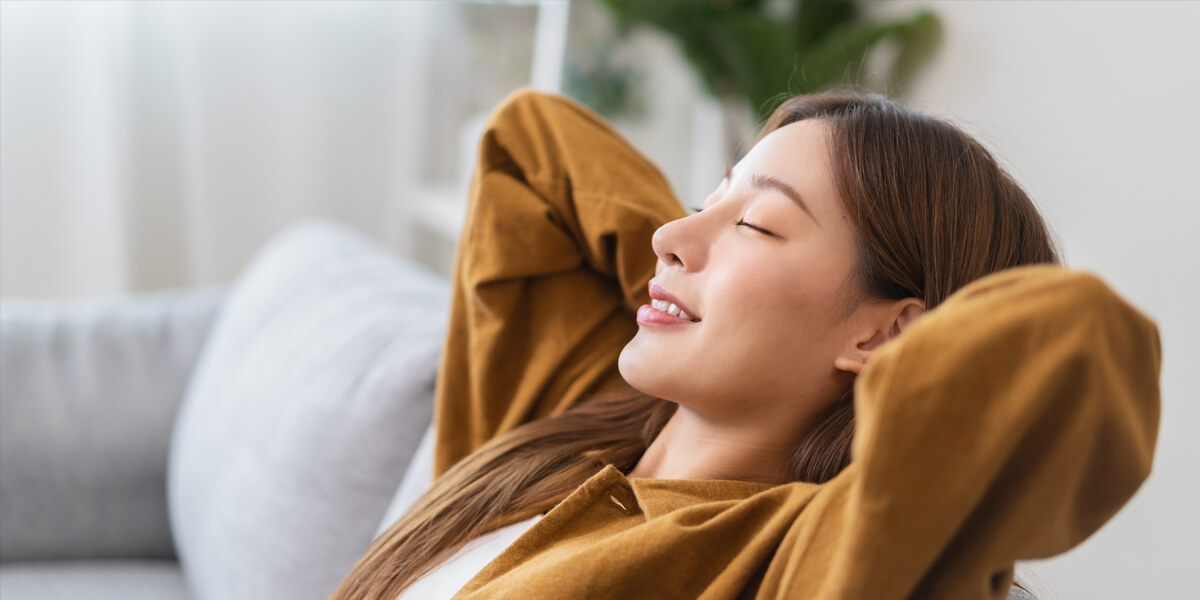 A woman with long hair relaxes on a couch, leaning back with her hands behind her head and eyes closed, smiling peacefully—perhaps unwinding after overseeing a roofing process. She is wearing a brown shirt. The background is softly blurred.