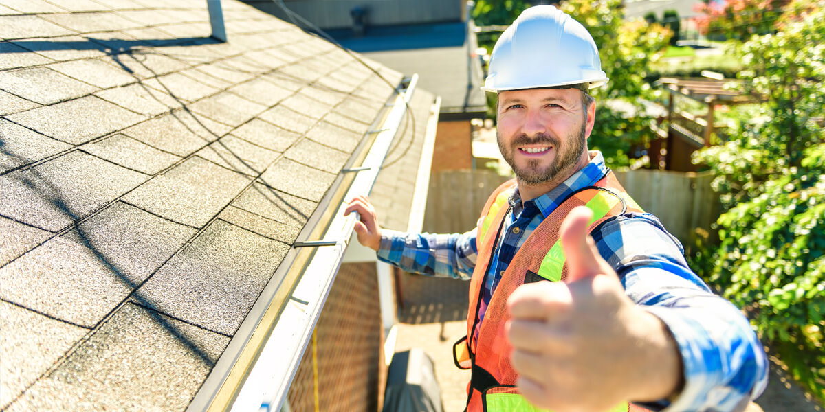 A construction worker wearing a hard hat and safety vest stands on a ladder next to a roof gutter, smiling and giving a thumbs-up to the camera, showcasing professional roofing Ottawa services on a sunny day.