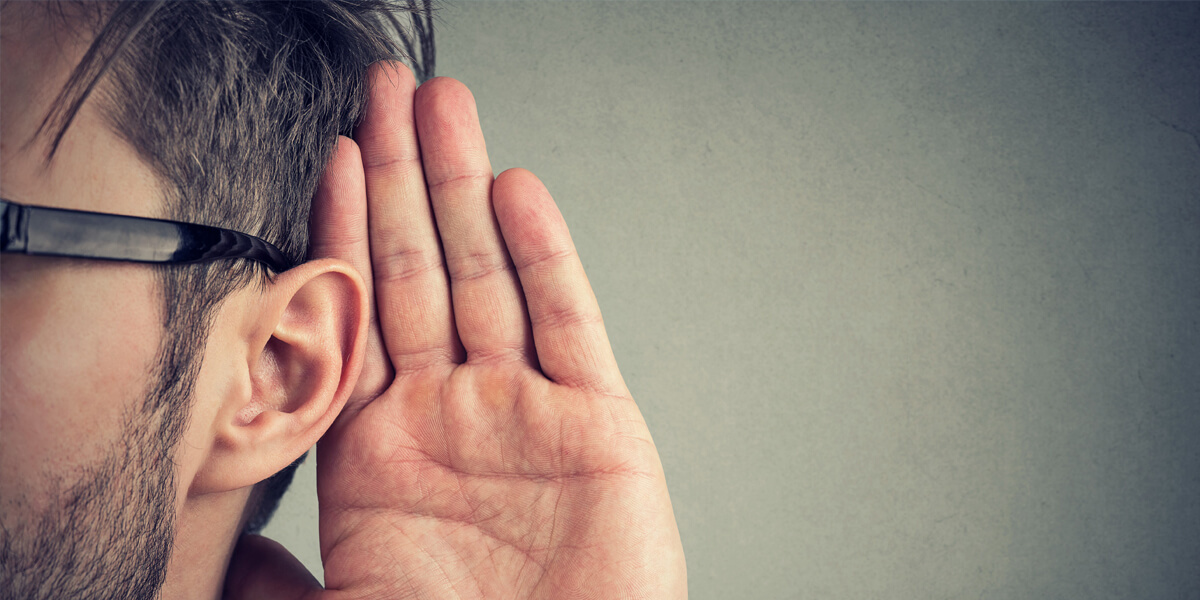 A person wearing glasses holds their hand up to their ear, appearing to listen closely. Only the side of the persons face and the hand are visible, with a neutral background.