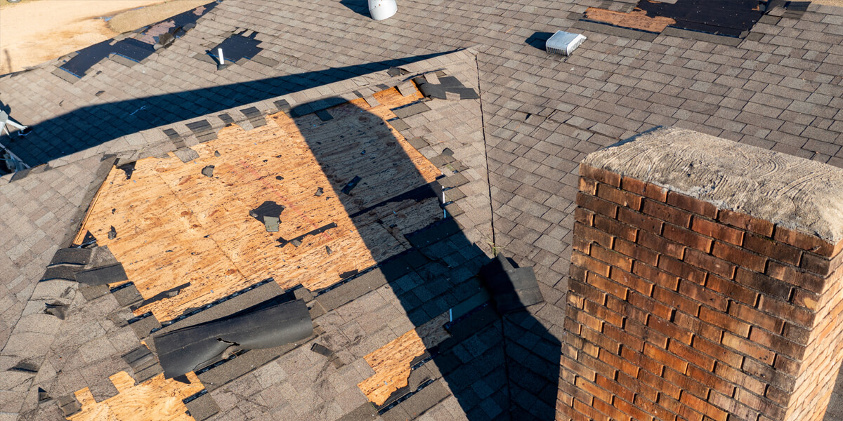 A roof with missing and damaged shingles exposes the wooden decking beneath. A brick chimney stands on the right, and scattered roofing materials suggest recent storm damage or repair work—an ideal situation to review your roofing insurance coverage.