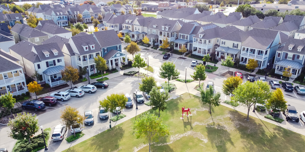 Aerial view of a suburban neighborhood with rows of houses featuring Stittsville roofing, parked cars along the street, and a small park area with green grass, trees, and a red playground structure.