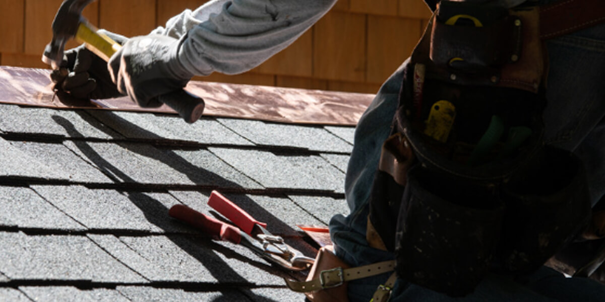 A worker wearing a tool belt uses a hammer to install or repair asphalt shingles on a roof beneath sunlight. The persons face is not visible, capturing the dedication seen in Riverside South roofing projects.