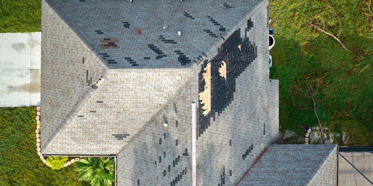 Aerial view of a house roof with missing and damaged shingles, revealing exposed areas in need of roof repairs Riverside South. The surrounding yard is green with grass and a few plants visible.