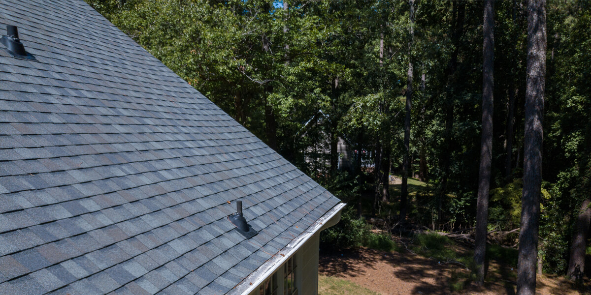 A roof features gray asphalt shingles and two vent pipes, viewed from above, with a grassy yard and trees in the background on a sunny day in an Orleans neighbourhood.