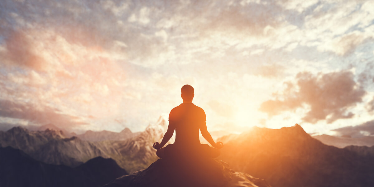 A person sits cross-legged on a mountaintop, meditating at sunrise or sunset, embracing peace of mind as the mountains and a colorful sky create a serene backdrop.