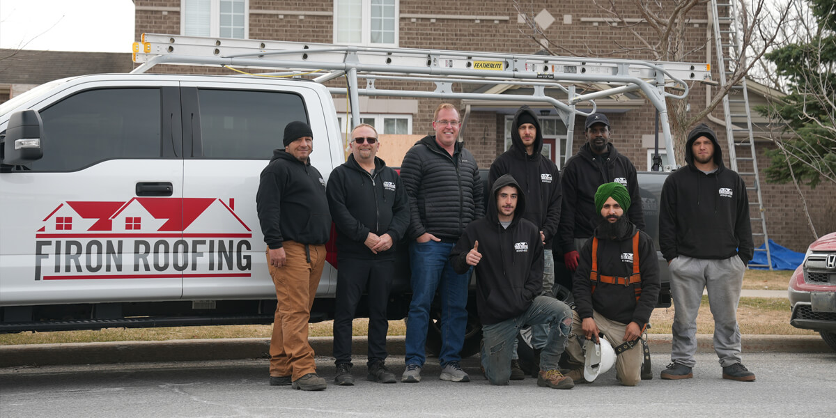 The Firon Roofing team in Ottawa, eight men in work attire, pose in front of a van with a ladder on top. Some stand while others kneel outdoors near houses, dressed in hoodies and jackets.