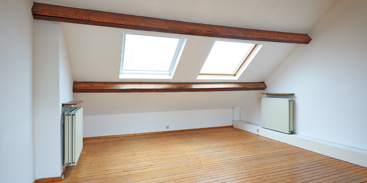 A bright attic room with wooden floors, white walls, exposed wooden beams, and two large skylights letting in natural light—ideal for a future skylight replacement. Two radiators sit on opposite walls beneath the sloped ceiling.