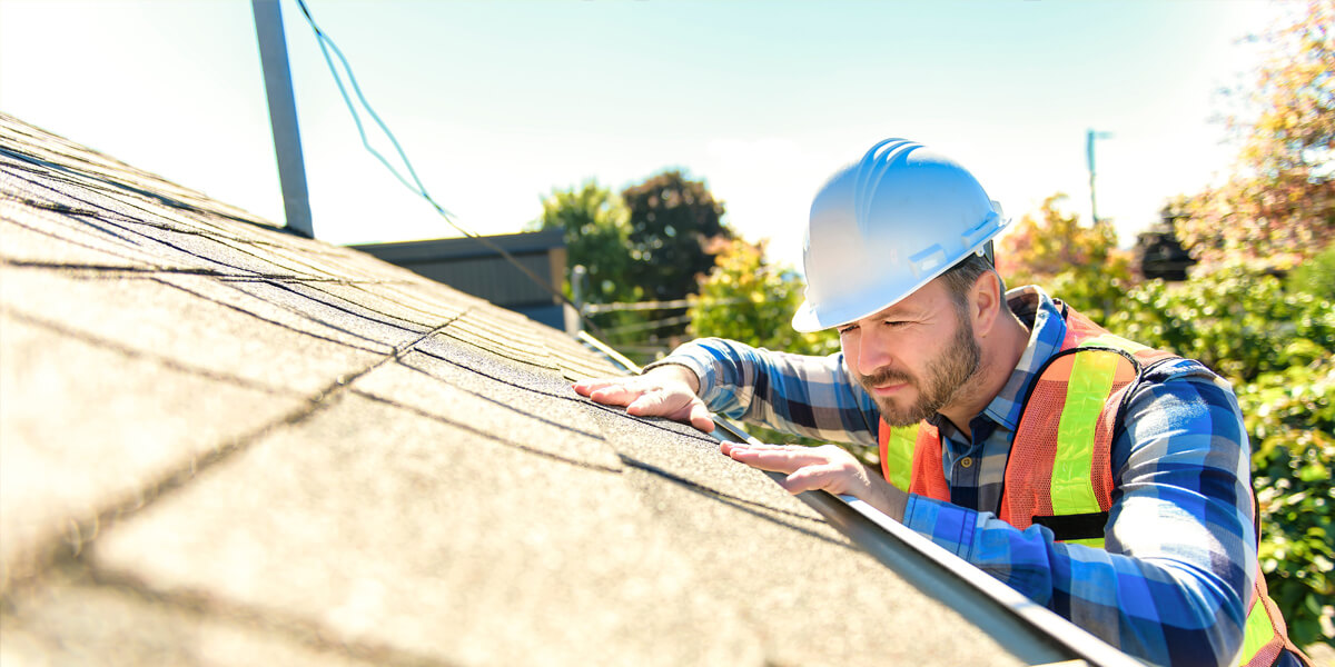 A construction worker wearing a white hard hat, safety vest, and plaid shirt inspects a residential roof under bright sunlight, checking the shingles for quality to ensure the use of the best roofing products. Trees and sky are visible in the background.