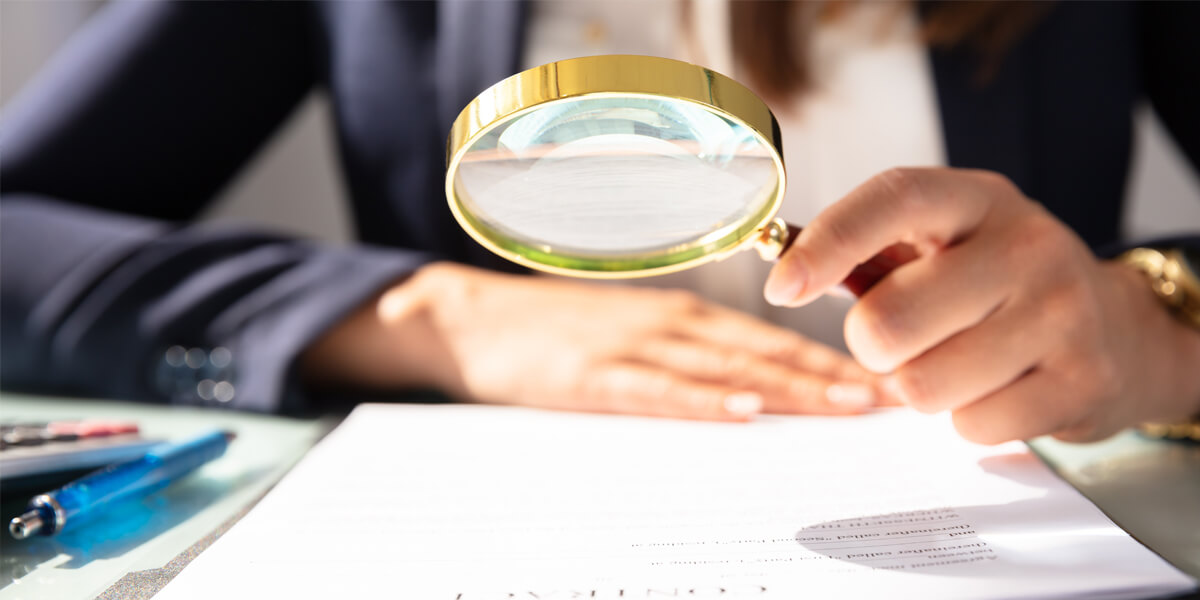 A person holding a magnifying glass over a document, closely examining the fine-print warranty. The individual is wearing a dark suit, and only their hands and partial torso are visible.