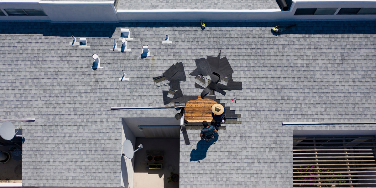 A person in a hat works on roof damage repairs, surrounded by scattered roofing materials and tools. The image, taken from above, shows a section of exposed wood where shingles have been removed.