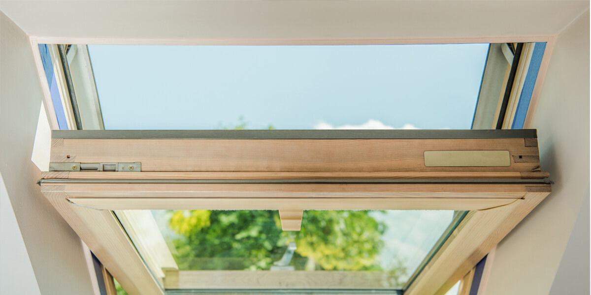 Open skylight window in a slanted roof, showing a clear blue sky and green trees outside. Sunlight streams through the glass, illuminating the wooden frame—a perfect example of quality skylights Nepean residents enjoy.