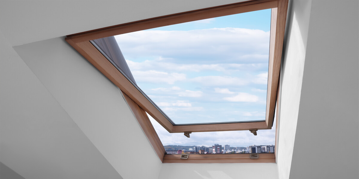 A skylight Nepean window set in a sloped white ceiling is open, revealing a view of a blue sky with clouds and a city skyline in the distance. The window has a wooden frame.