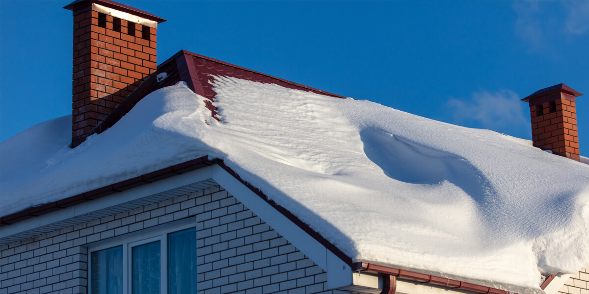 A house with white brick walls and a red roof covered in thick snow, with a red brick chimney and blue sky in the background—perfectly capturing a scene before roof snow removal Ottawa services are needed. Snow piles up on the sloped roof above a window.