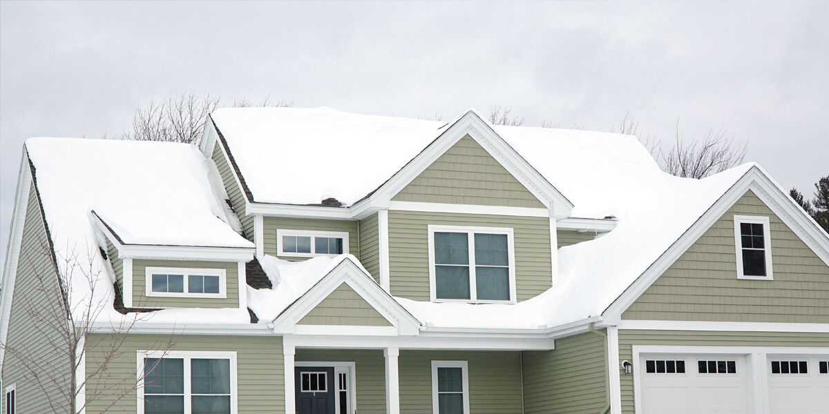 A green two-story house with white trim sits under a cloudy winter sky, its roof and yard blanketed in snow—an ideal candidate for professional snow removal Manotick services.