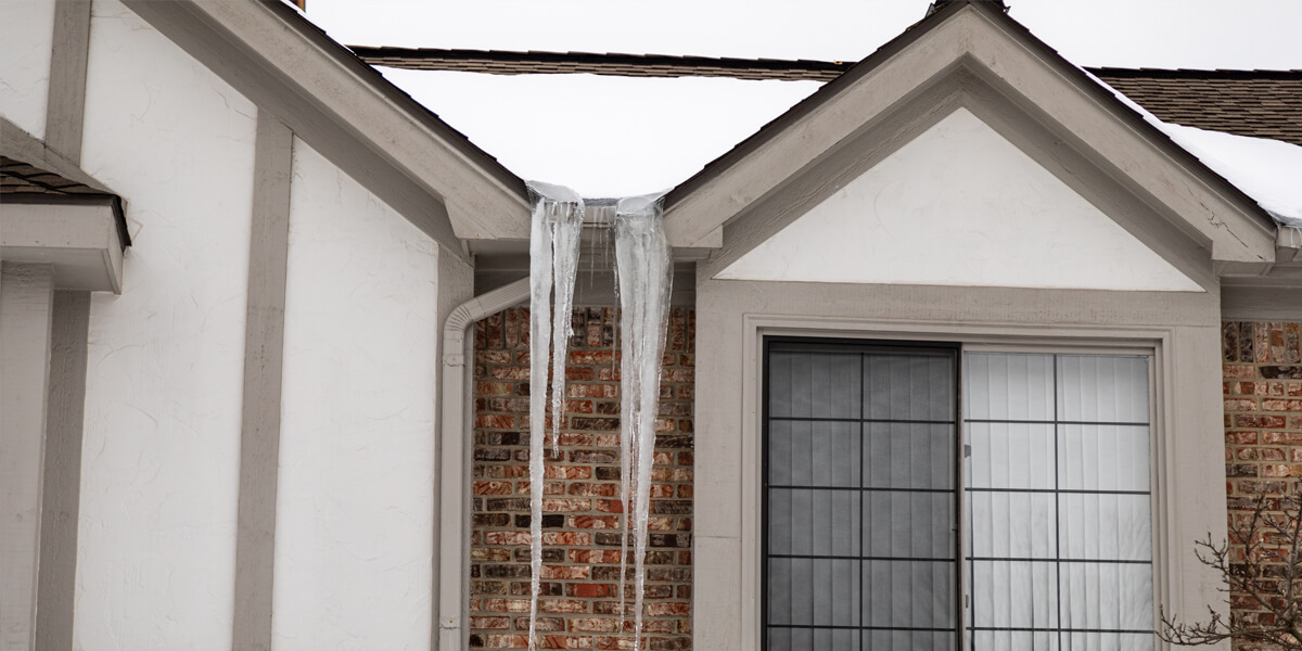 Two large icicles hang from the gutter of a brick house with snow on the roof and ground—an all-too-common sight before ice dam removal in Manotick. A window with closed blinds is visible beneath the thick icicles.