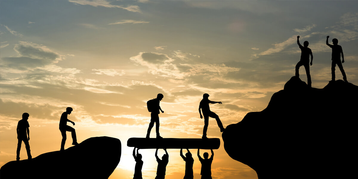 Silhouettes of people at sunset, some holding up a beam to form a bridge while others walk across or prepare to step onto it, symbolizing teamwork and cooperation.