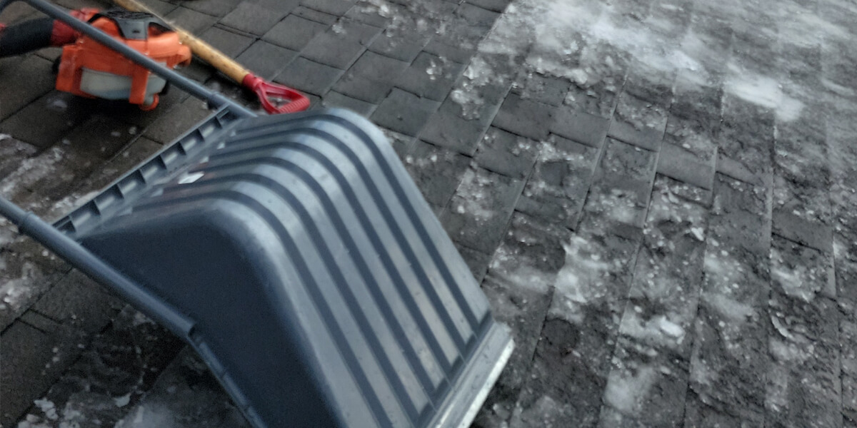 A snow-covered roof with patches of ice—ideal for ice dam removal in Manotick—a snow shovel and an orange ice chopper rest on the shingles near a brick house in winter.
