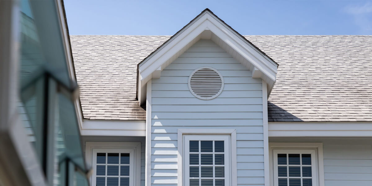 Close-up of a light blue house exterior with white trim, featuring a gable roof, a round attic vent, and two windows under a clear blue sky—ideal for showcasing roof inspection Ottawa services.