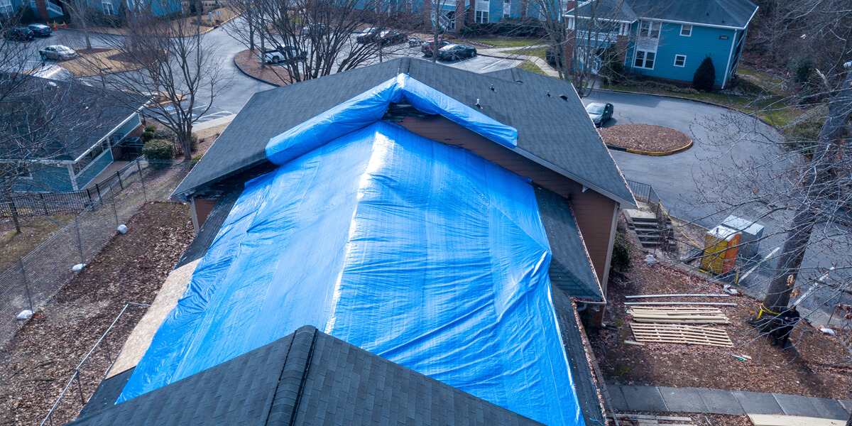 Aerial view of a house with a large blue tarp covering the roof, likely due to roof wind damage in Kanata, surrounded by trees and neighboring homes. Construction materials are visible on the ground near the building.