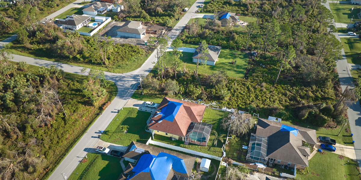 Aerial view of suburban houses in Kanata, Ontario, some with blue tarps covering storm-damaged roofs. Green lawns and trees surround the homes while roads divide the neighborhood.