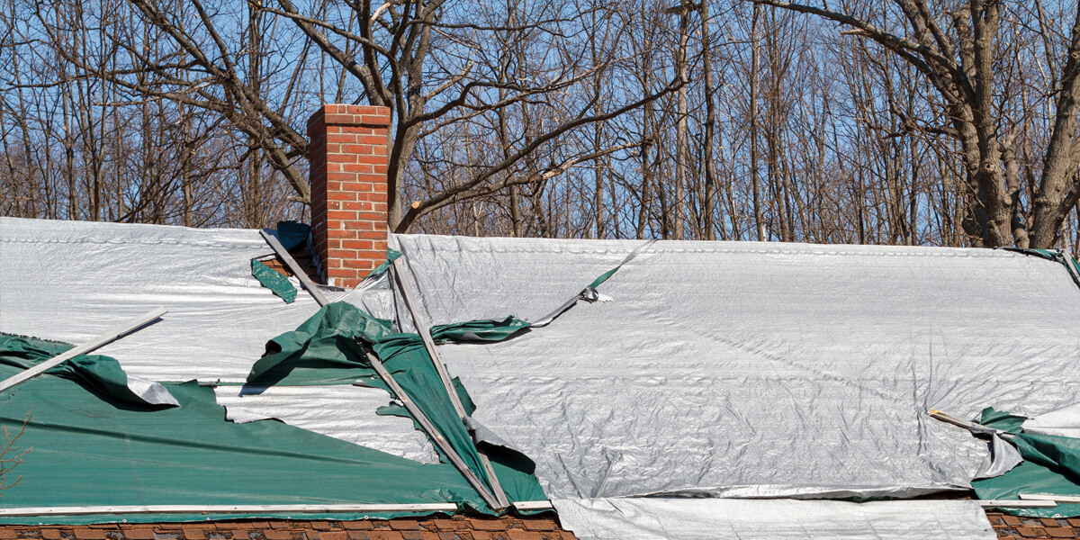 A Greely Ontario roof with damaged tarps flapping in the wind exposes shingles beneath. A brick chimney rises at the center, framed by leafless trees in the background.