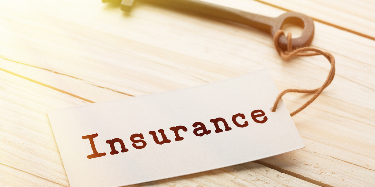 A vintage key rests on a wooden surface beside a white tag labeled Insurance in bold, brown letters. The tag, attached with string, hints at the importance of roof insurance Greely for safeguarding cherished possessions.