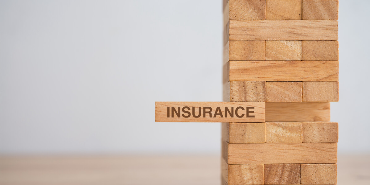 A Jenga tower made of wooden blocks, with one block labeled Greely insurance being pulled out, suggesting risk or instability without Greely insurance.