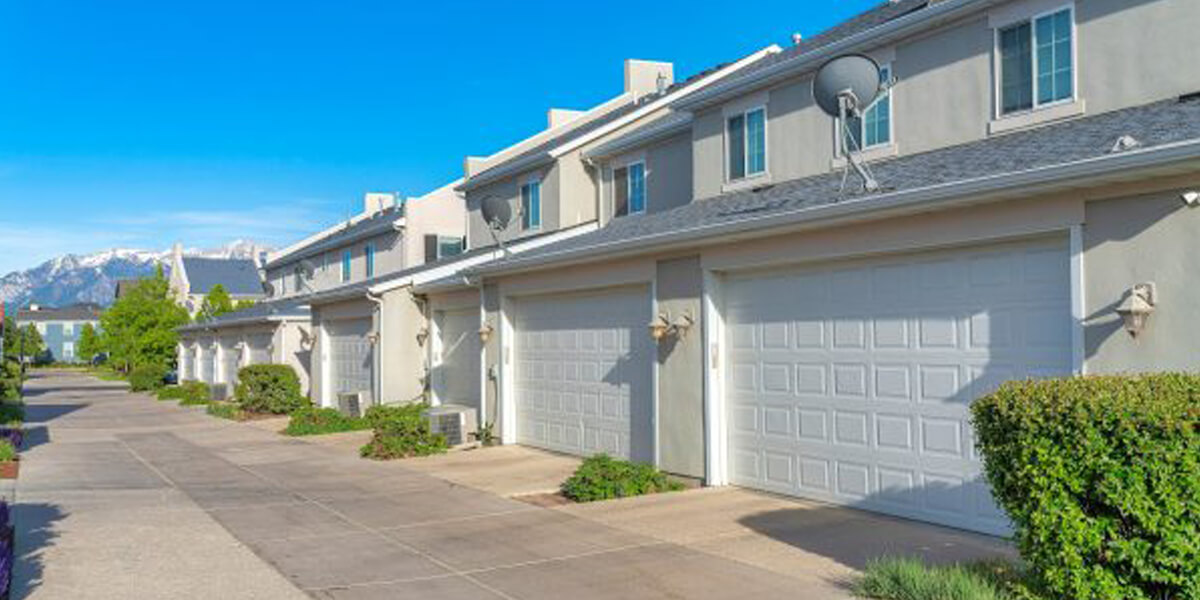 A row of modern, beige condos in The Glebe with double garages and satellite dishes on the roof, lined along a clean, paved driveway under a clear blue sky. Shrubs neatly border each unit.