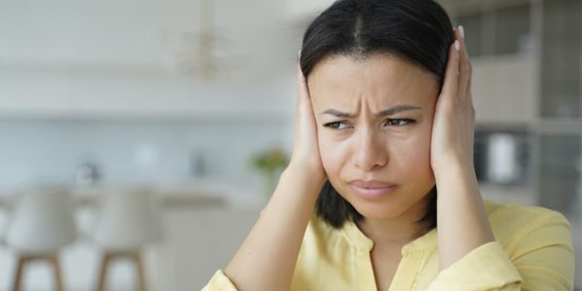 A woman in a yellow shirt covers her ears with her hands and looks upset, possibly reacting to loud roofing behaviours, sitting in a bright, blurry indoor setting.