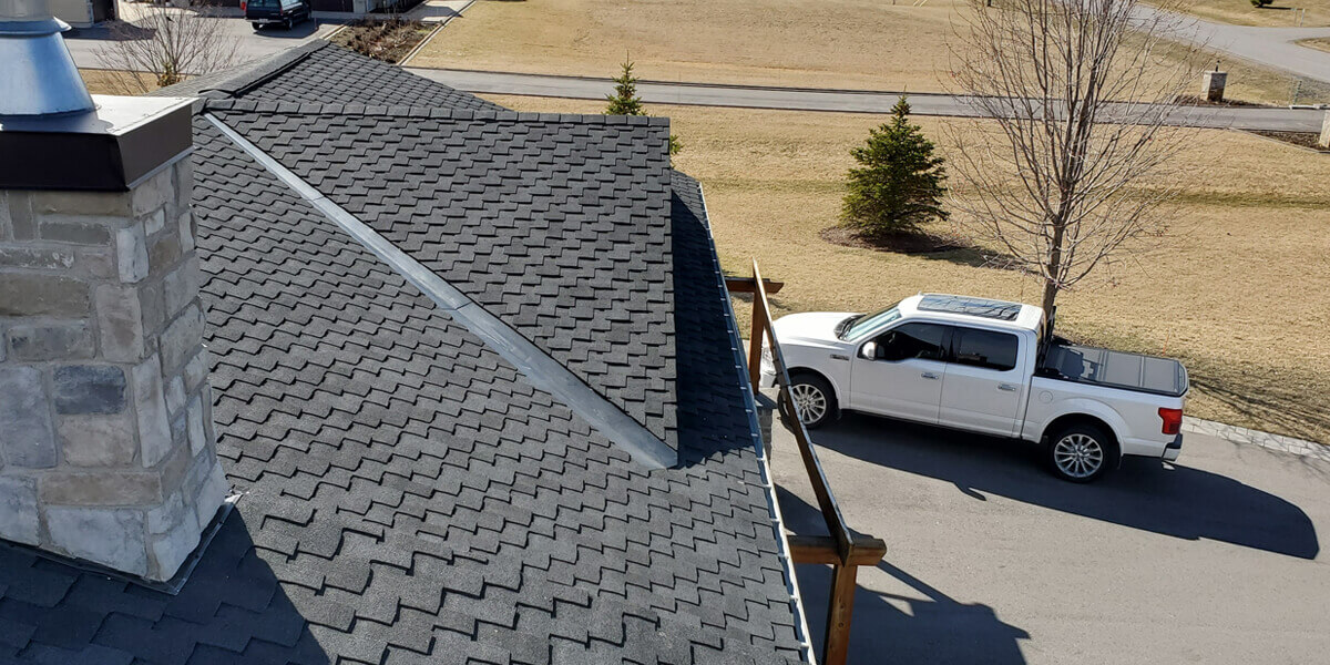 A white pickup truck is parked on a driveway beside a house with dark gray asphalt shingles, possibly after a roof replacement Ottawa homeowners might choose. The yard features dry grass, some small evergreens, and a leafless tree near the driveway.