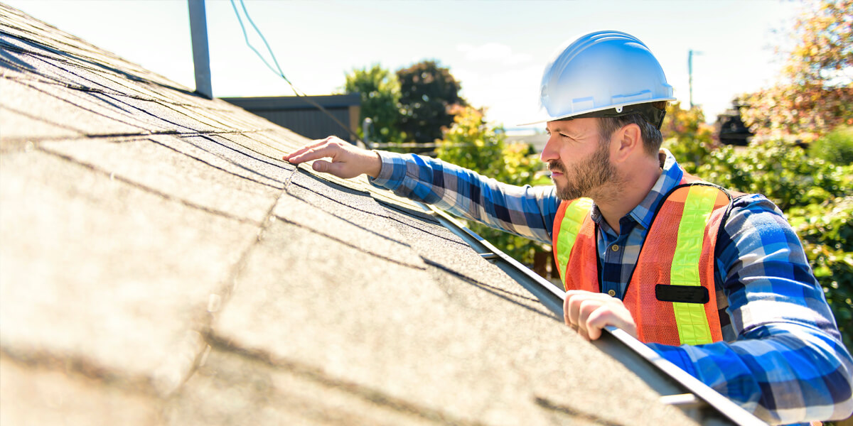 A construction worker from an Ottawa roofing company, wearing a hard hat, safety vest, and plaid shirt, inspects the shingles on a residential roof on a sunny day, surrounded by trees in the background.