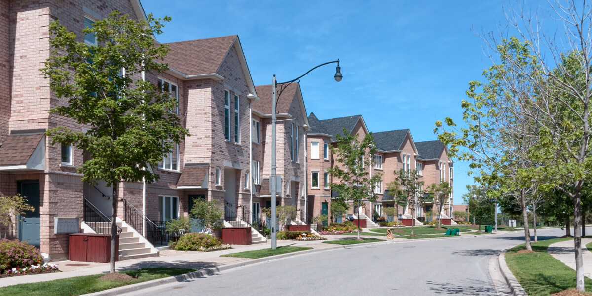 A quiet suburban street lined with modern, two-story brick houses featuring quality roofing in the Glebe, green lawns, and young trees under a clear blue sky.