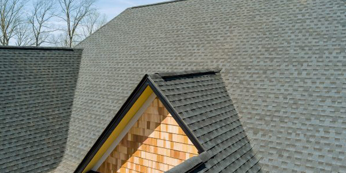 Close-up view of a gray shingled roofing Barrhaven home with a triangular dormer featuring tan wooden siding, set against a clear sky and bare tree branches in the background.