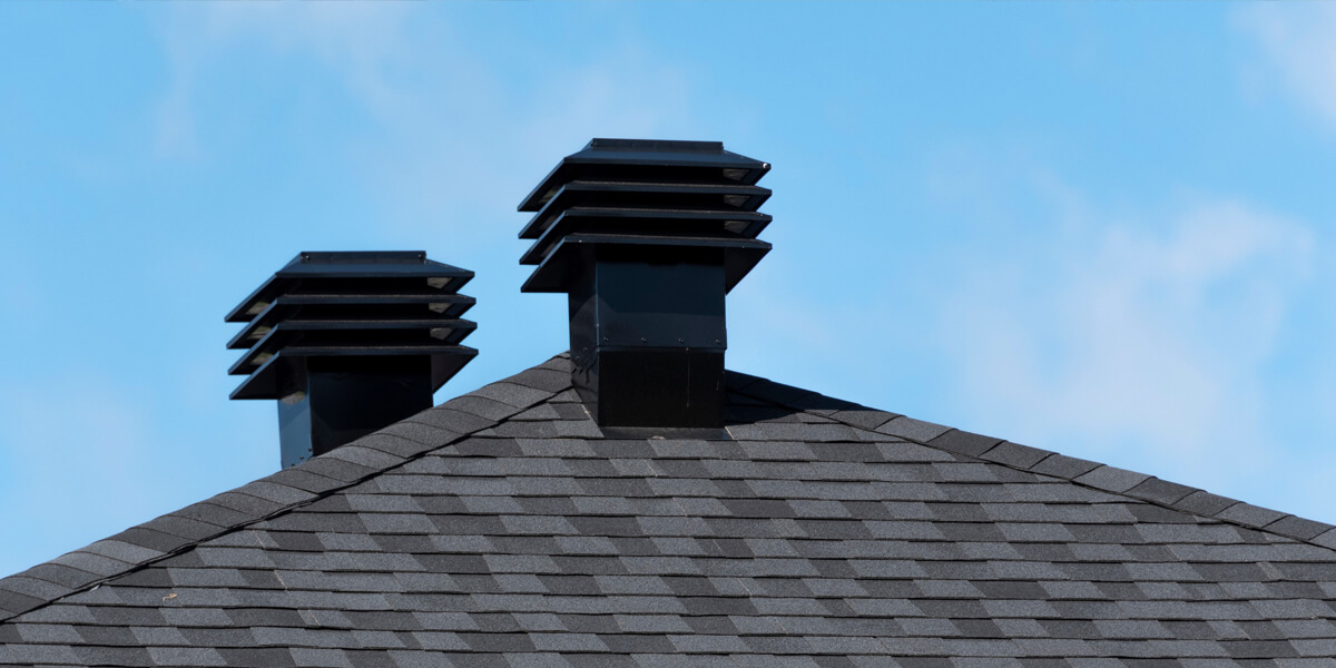 Two square black roof vents with horizontal slats on a gray shingled roof, set against a clear blue sky.