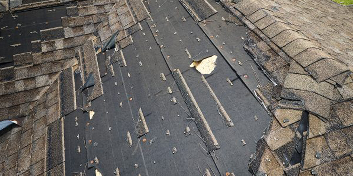 A roof with missing and damaged shingles exposes the black underlayment beneath. Some shingles are loose or curled up, and nails are visible, suggesting significant roof damage that may require a fiberglass roof replacement.