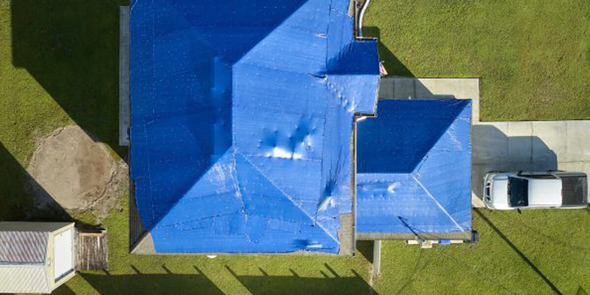 Aerial view of an Ottawa roof covered with a large blue tarp, surrounded by green grass, with a white car in the driveway. The tarp spans the entire roof, suggesting recent damage or repair work.
