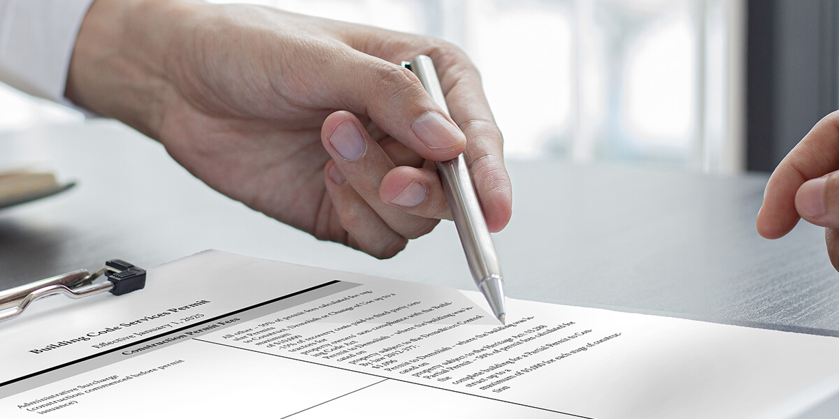 A Building Code Services roof permit Ottawa form with two pens and a paperclip is overlaid on an aerial view of residential buildings and a tree-lined street in autumn.