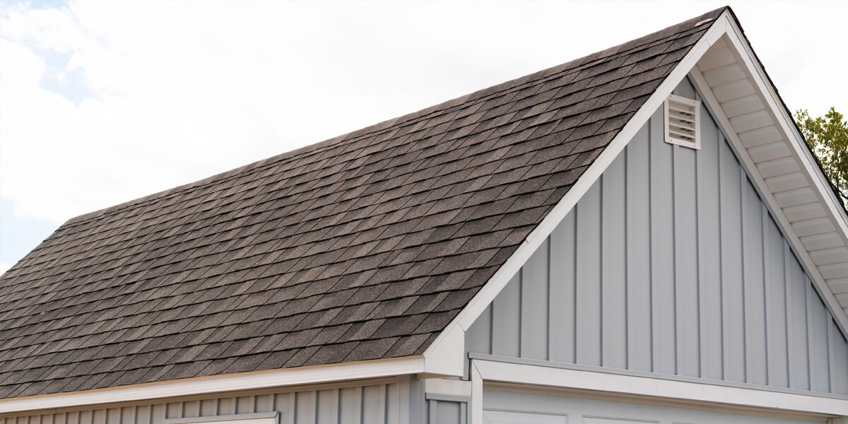 A close-up of a house with light blue vertical siding and a dark gray shingled roof, showcasing expert roof replacement Ottawa. There is a white vent near the roof peak and white trim along the edges.