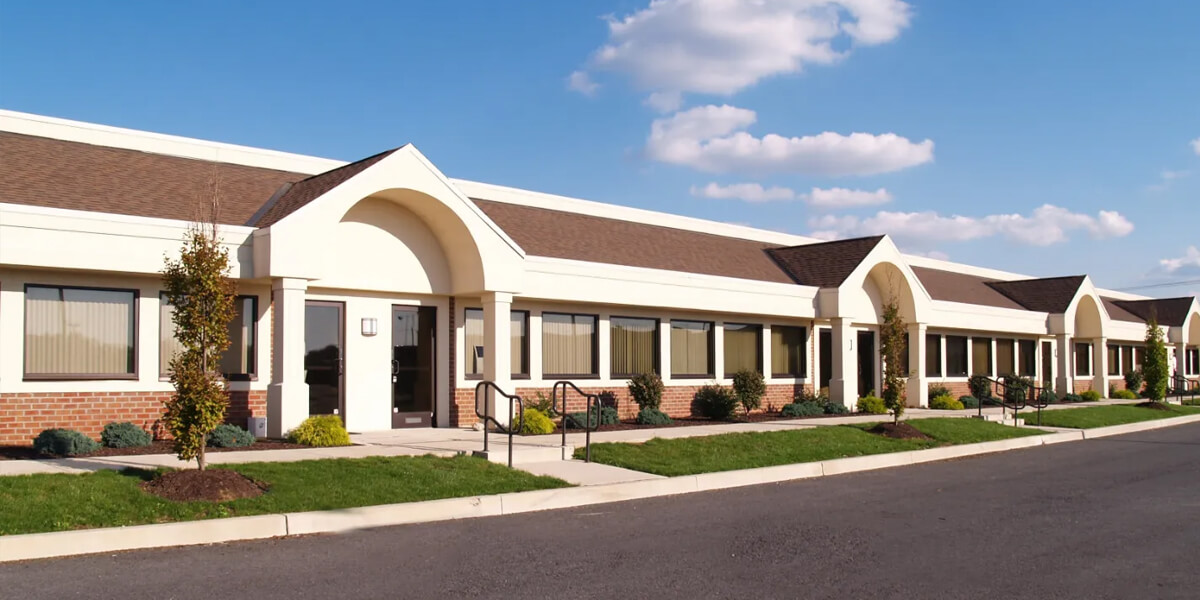 A single-story office building with large windows, brick and cream-colored facade, arched entryways, landscaped shrubs, and a paved parking lot—ideal for businesses seeking quality B2B roofing solutions—stands under a blue sky with scattered clouds.