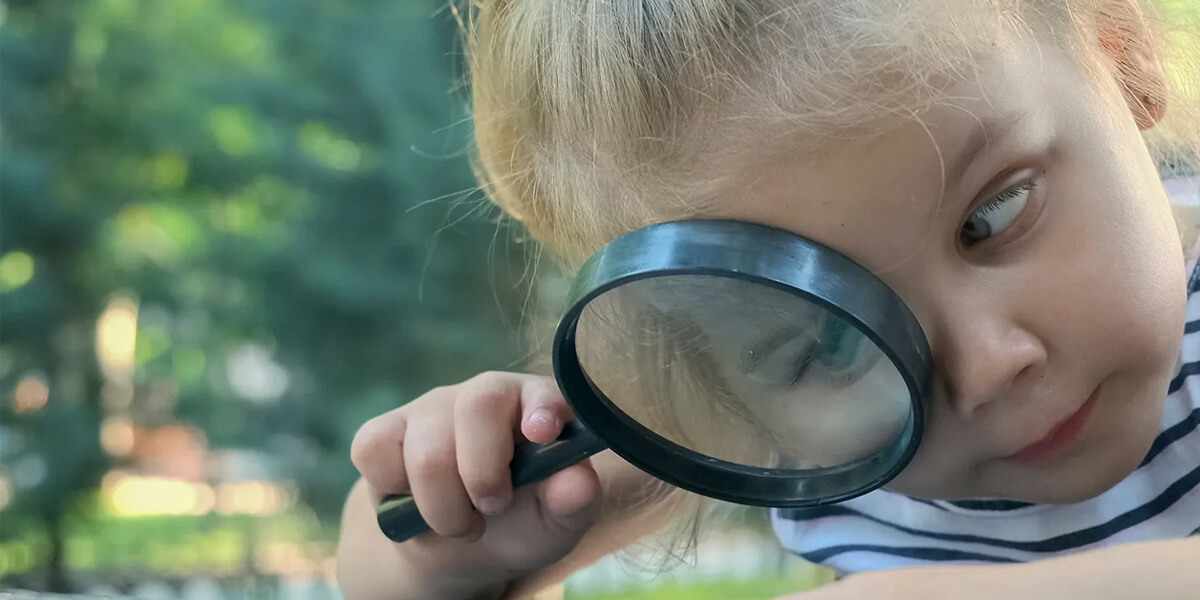 A young child with blonde hair looks through a magnifying glass, examining roofing details closely outdoors. The child is wearing a striped shirt and gazes to the side with curiosity.