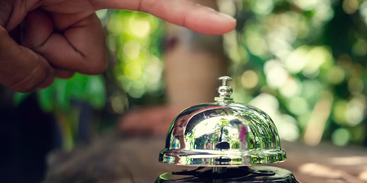A close-up of a hand about to press a shiny silver service bell on a wooden surface, with a blurred green background.