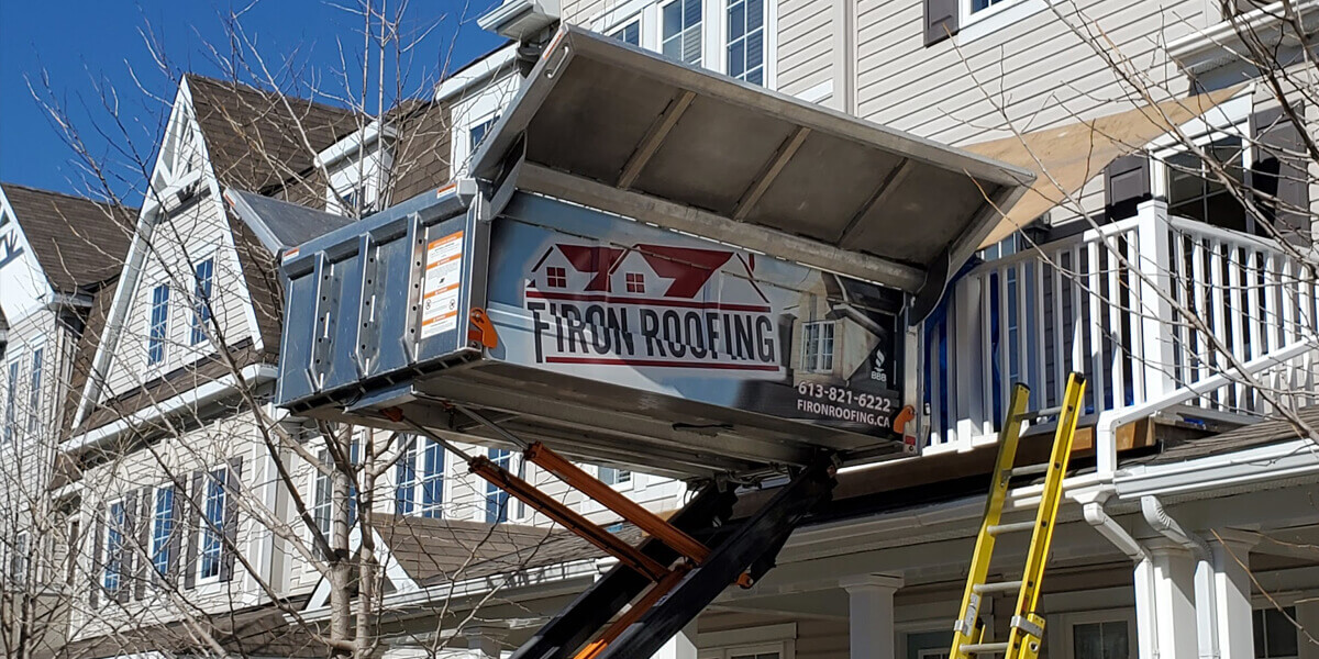 A metal dumpster labeled Firon Roofing is lifted by a hoist to an upper floor of a beige house with white trim. A yellow ladder leans against the porch, and bare tree branches are visible.