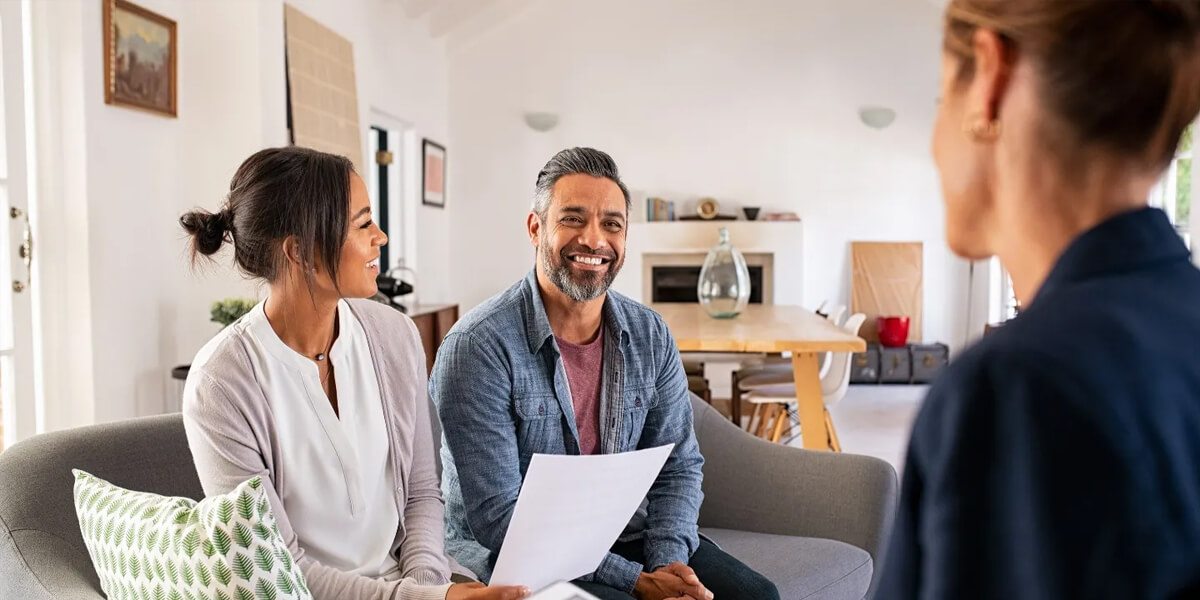 A smiling couple sits on a couch holding papers, talking to a woman whose back is to the camera, in a bright, modern living room with a dining table in the background.