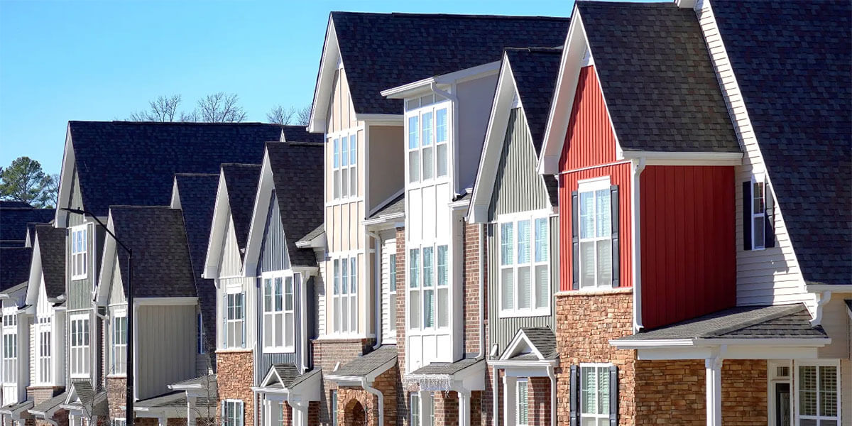 A row of modern, multi-story townhouses with varying colors of siding and brick exteriors, sharply peaked roofs featuring quality roofing for property managers, and large windows under a clear blue sky.