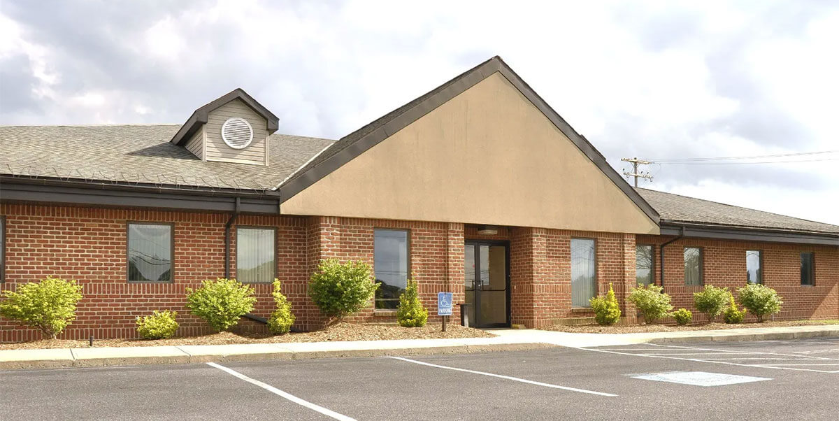 A single-story brick office building with large windows and a peaked roof, surrounded by small green shrubs and an empty parking lot in front—ideal for property owners Ottawa seeking an attractive, low-maintenance space.