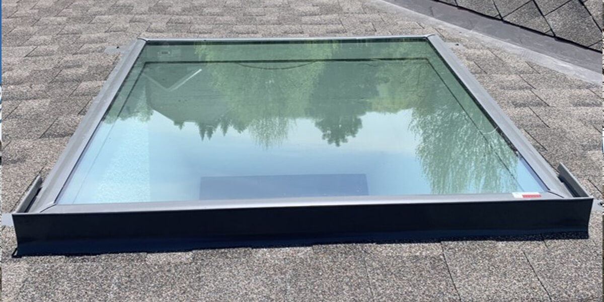 A rectangular skylight with a metal frame is installed on a shingled rooftop, reflecting trees and the sky above—an elegant example of skylights for roofs enhancing natural light.