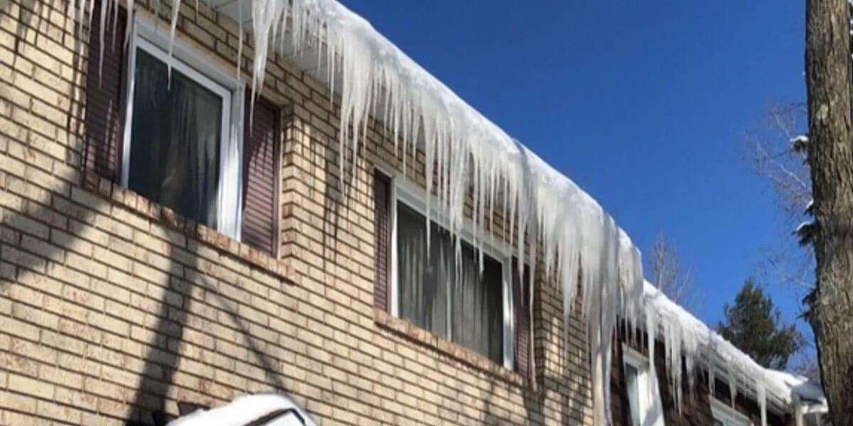 A two-story brick house with long, sharp icicles hanging from the roof and closed blinds on the windows under a clear blue sky. Snow covers the lower roof, highlighting the need for timely roof snow and ice removal.