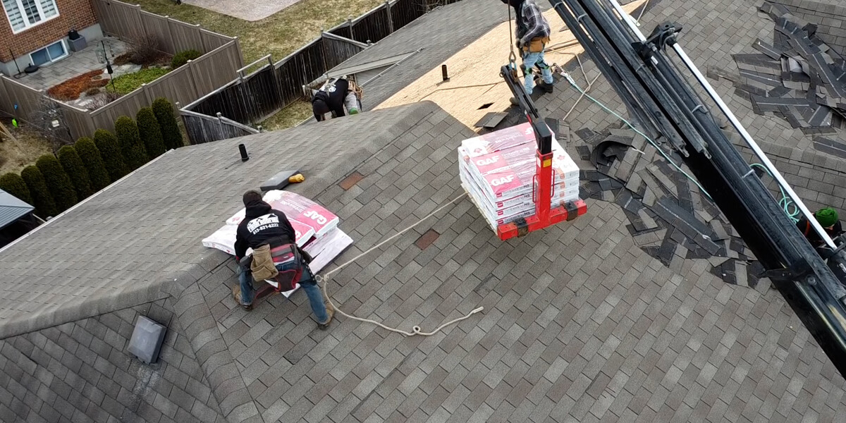 A worker installs shingles on a sloped roof while a crane lifts bundles of roofing shingles onto the roof. Other workers and materials are visible, showcasing the expertise of top roofers in North America in a residential neighborhood.