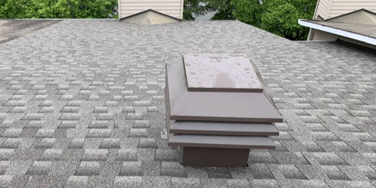 A brown roof vent, part of recent roof ventilation upgrades, sits on a gray shingled roof dotted with water droplets. In the background, trees and neighboring buildings with beige siding and similar vents are visible.